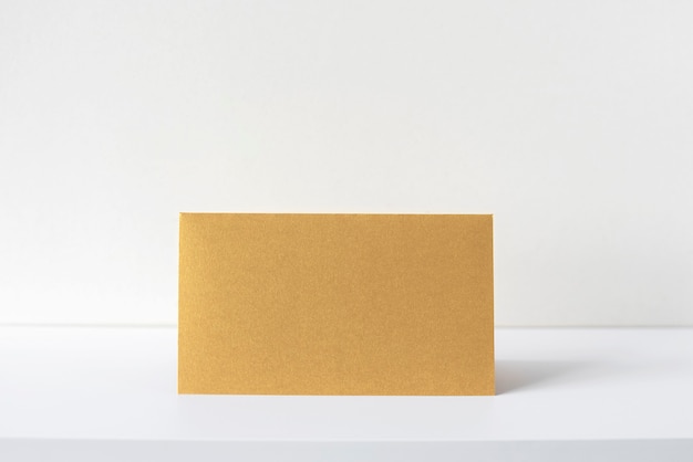 Goldene Einladung oder Grußkarte auf weißem Schreibtisch, beige Interieur. Leeres quadratisches Papierblatt auf dem leeren Tisch. Mockup, Platz für Text.