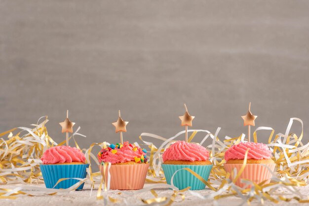 Goldene Kerzen in Sternform auf Cupcakes. Muffins mit rosa Buttercreme-Zuckerguss auf festlichem Lametta-Hintergrund. Kopieren Sie spacve auf grauem Hintergrund.