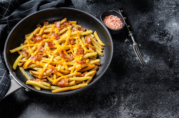 Goldene Pommes Frites mit Cheddar-Käse und Speck in einer Pfanne gebraten. Schwarzer Hintergrund.