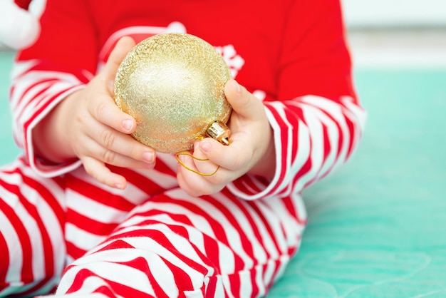 Goldener Weihnachtsball in den Händen eines kleinen Babys, das Weihnachtskostüm trägt, Nahaufnahme neues Jahr und