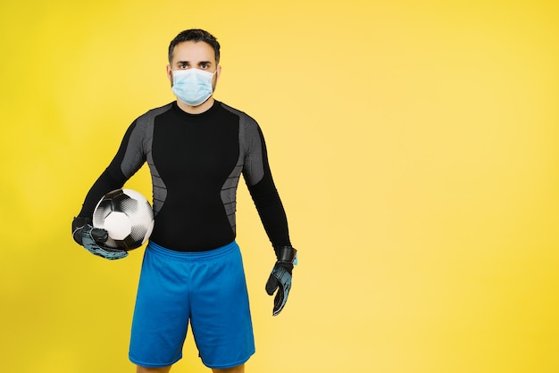 Foto goleiro de futebol com máscara no rosto devido à pandemia de coronavírus covid19 com a bola com as mãos na parede amarela