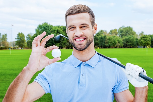 Golfista exitoso. Feliz joven sosteniendo la pelota de golf y el conductor mientras está de pie en verde