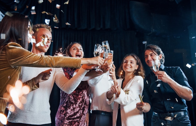 Golpeando vasos. Grupo de amigos alegres celebrando el año nuevo en el interior con bebidas en las manos.