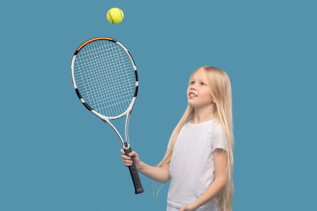 Golpear la bola. Chica enérgica alegre en camiseta blanca de deportes en deleite golpeando la pelota con raqueta de tenis