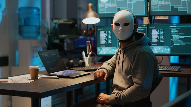 Golpista mascarado hackeando rede de computadores tarde da noite, trabalhando em laptop e vários monitores para roubar informações de TI. Hacker com servidor de segurança de quebra de identidade oculta, ataque cibernético.