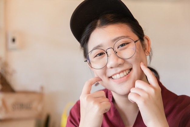 Gordita rechoncha adolescente linda sonrisa de diente blanco Joven estudiante asiática con gafas y sombrero dedo toque mejilla con espacio de copia