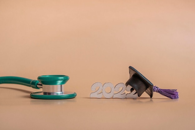 Gorra de graduación con estetoscopio y números 2022 sobre fondo beige, primer plano con espacio de copia. Concepto de educación médica.