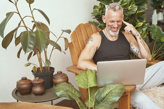 Foto gostando de trabalhar em casa, feliz homem caucasiano de meia-idade usando laptop e sorrindo enquanto está sentado