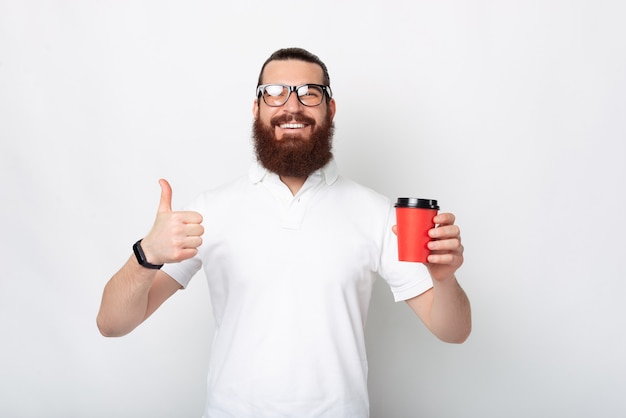 Gosto de levar meu café comigo. Homem barbudo segurando um copo de papel vermelho e aparecendo o polegar.