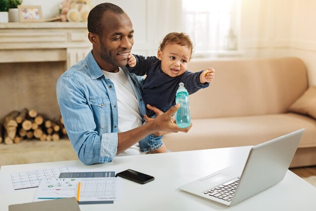 Gostoso. Sorrindo, amoroso jovem pai afro-americano segurando seu filho pequeno e alimentando-o enquanto está sentado à mesa com seu laptop