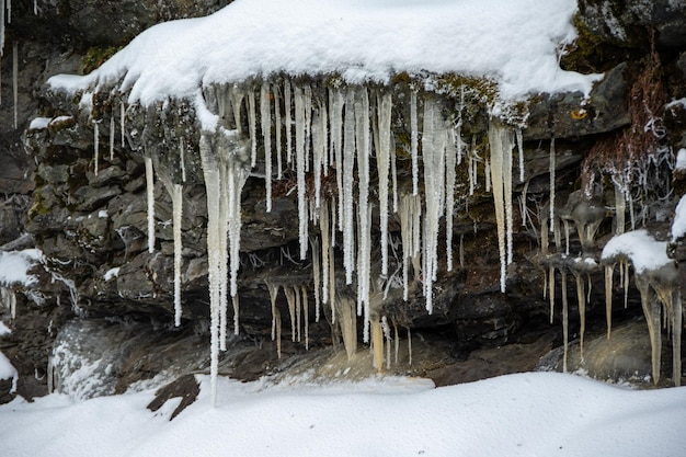 Gotas de água derretida do telhado caem na neve e pingentes de gelo congelados ao lado de pedra na primavera pingentes de gelo e um jato de água