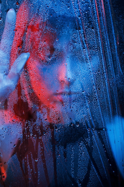 Gotas de água. Estúdio filmado em estúdio escuro com luz de néon. Retrato de uma linda garota atrás de um vidro molhado