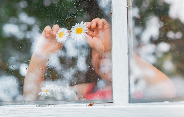 Gotas de lluvia en el cristal de la ventana de una aldea, flores de manzanilla, ojos en manos de niños, miran la lluvia.