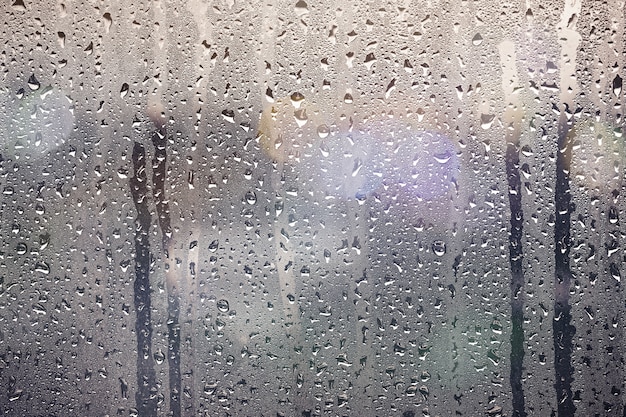 Gotas de lluvia en la ventana de cristal con el bokeh ligero de la noche cerca para arriba para el fondo