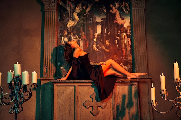 Goth-Thema Schöne junge Dame im kurzen schwarzen Kleid sitzt auf einem Steinsockel in der Nähe von Kerzen auf dunklem Hintergrund