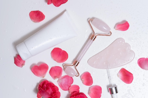 Gouache-Massagegeräte und Gesichtscreme liegen auf einem weißen Hintergrund mit Rosenblättern