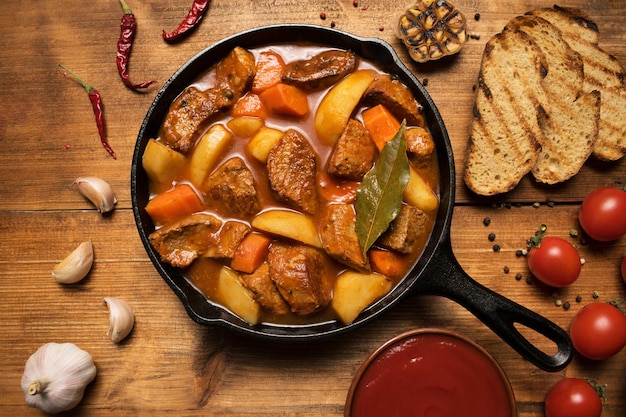 Goulash estofado de carne con patatas, zanahorias y verduras en una sartén de hierro fundido sobre un fondo de madera con picatostes, salsa, ajo y tomates. Cierre plano. Vista superior.