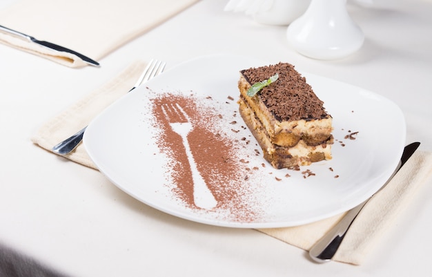 Gourmet-Scheibe leckeren Schokoladenkuchen auf weißem Teller mit Gabel-Umriss mit Kakaopulver auf der Seite. Serviert auf weißem Esstisch.