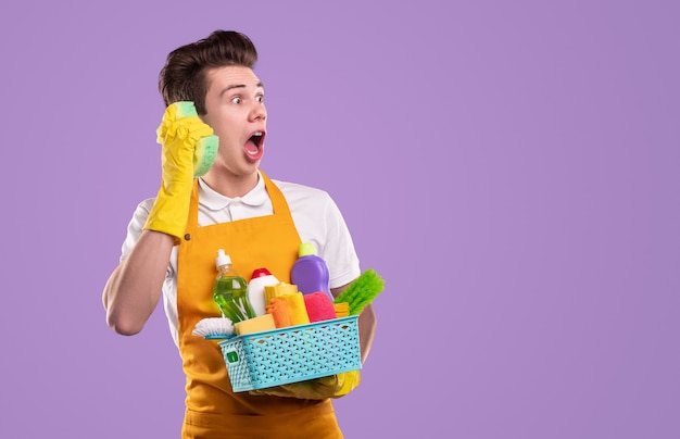 Governanta louca com um recipiente de vários detergentes segurando uma esponja perto da orelha como um telefone e olhando para o outro lado com espanto contra um fundo violeta