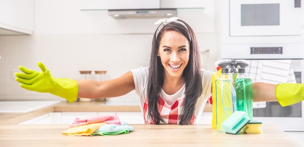 Governanta sorridente com os braços bem abertos na cozinha, com trapos coloridos e detergentes sobre a mesa.