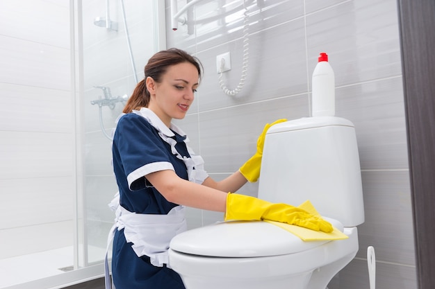 Governanta trabalhadora em um avental branco ajoelhada limpando um assento de vaso sanitário limpando a tampa com um pano