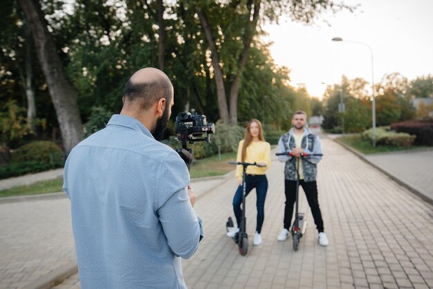 Graban un video blog sobre una joven pareja hermosa que monta patinetes eléctricos en un parque en un cálido día de otoño. Pasatiempos y recreación.