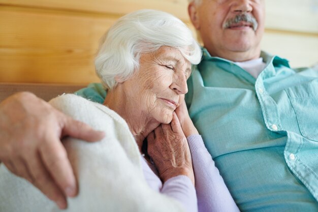 Graciosa abuela con cabello blanco tomando una siesta en el hombro de su esposo mientras ambos se relajan en el sofá frente al televisor