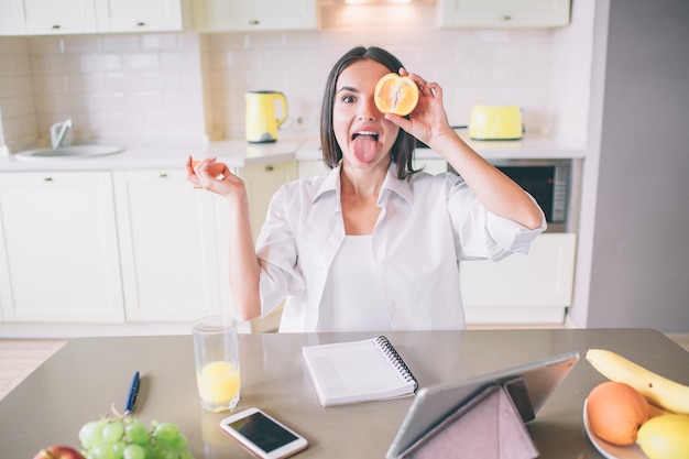 Graciosa está jugando con fruta. Sostiene un trozo de naranja delante del ojo y muestra su lengua.