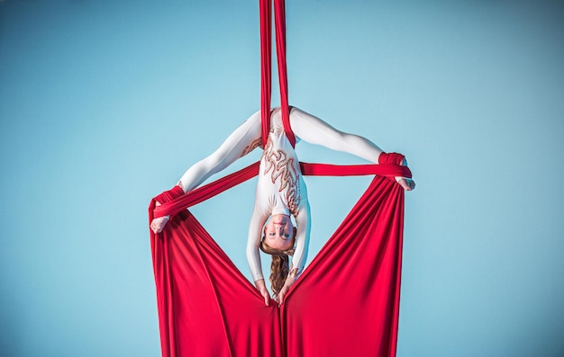 Graciosa ginasta fazendo exercícios aéreos com tecidos vermelhos sobre fundo azul