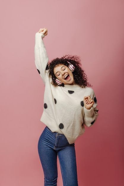 Graciosa joven caucásica con auriculares cerrando los ojos posando en el interior Bonita mujer morena con suéter y jeans escucha música Concepto de disfrutar el momento