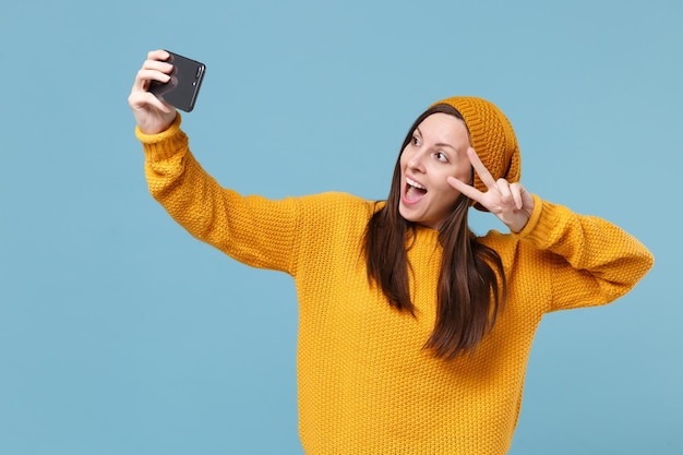 Graciosa joven morena con sombrero de suéter posando aislada en un retrato de estudio de fondo azul. Concepto de estilo de vida de las personas. Simulacros de espacio de copia. Haciendo un selfie en un teléfono móvil mostrando el signo de la victoria.