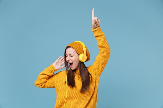 Graciosa joven morena con sombrero de suéter posando aislada en un retrato de estudio de fondo azul. Gente emociones sinceras concepto de estilo de vida. Simulacros de espacio de copia. Escucha música con auriculares bailando.