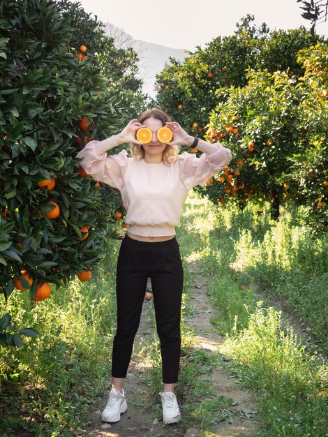 Graciosa mujer bonita joven de pie en el huerto de naranjos y sosteniendo dos mitades de naranja delante de sus ojos