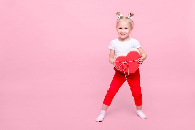 Graciosa niña en una camiseta blanca y pantalón rojo con una caja en forma de corazón sobre un fondo rosa. Retrato infantil con espacio para texto.