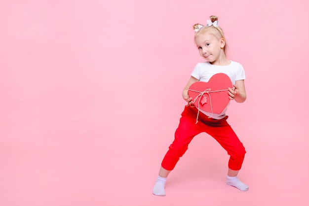 Graciosa niña en una camiseta blanca y pantalón rojo con una caja en forma de corazón sobre un fondo rosa. Retrato infantil con espacio para texto.