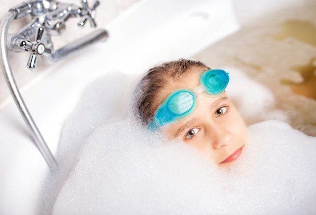 Graciosa niña caucásica positiva con gafas de natación y juega en el baño con espuma mientras espera la relajación junto al mar