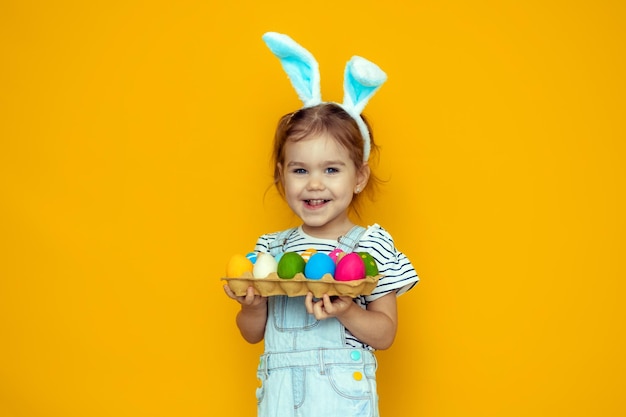 Graciosa niña feliz con huevos de pascua y orejas de conejo sobre fondo amarillo