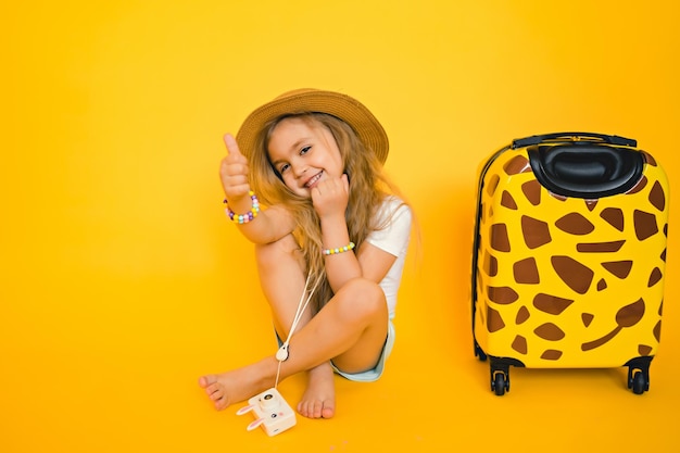 Foto graciosa niña sonriente con maleta amarilla esperando vacaciones levantando los pulgares