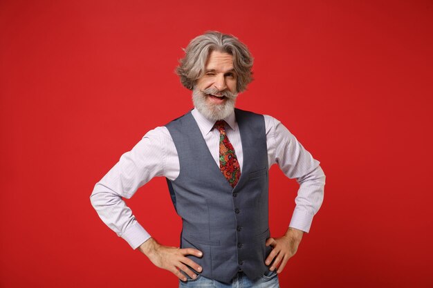 Gracioso anciano canoso bigote barbudo hombre en camisa clásica chaleco colorido corbata posando aislado sobre fondo rojo. Concepto de estilo de vida de las personas. Simulacros de espacio de copia. De pie con los brazos en jarras, parpadeando.