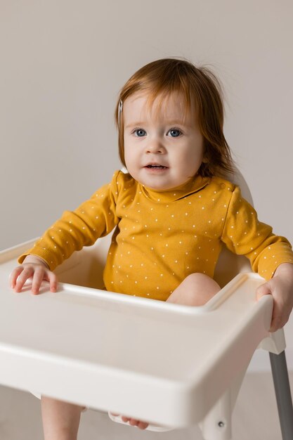 Gracioso bebé pelirrojo de ojos azules con un traje amarillo sentado en una silla alta blanca
