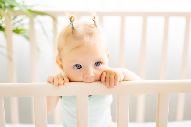 Gracioso bebé de pie en una cuna en una guardería blanca con una ventana Una niña pequeña en un traje de algodón Sonriente bebé de ojos azules