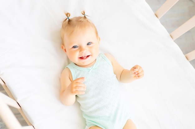 Gracioso bebé de pie en una cuna en una guardería blanca con una ventana Una niña pequeña en un traje de algodón Sonriente bebé de ojos azules