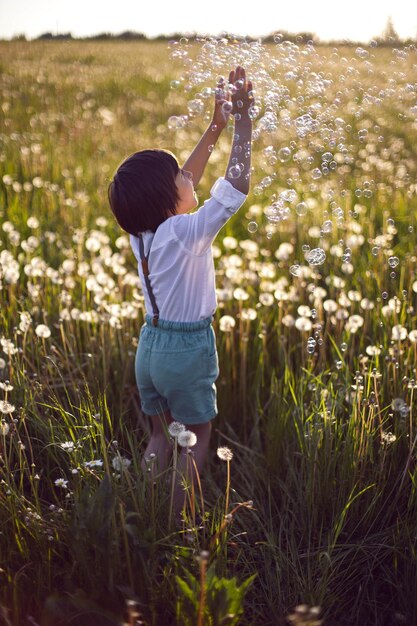 Gracioso, feliz, un hermoso niño se para en un campo con dientes de león blancos al atardecer en verano, las burbujas de jabón están volando