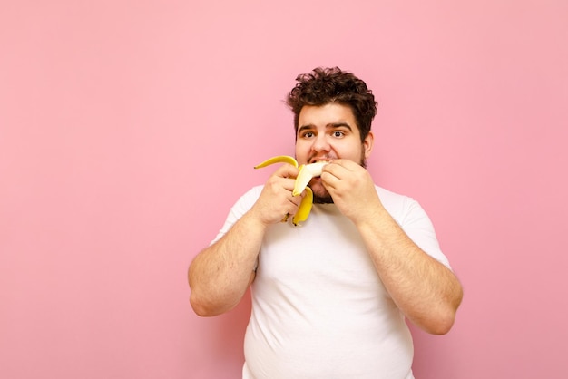 Gracioso gordo joven en camiseta blanca comiendo un plátano en una dieta aislado sobre un fondo de color rosa