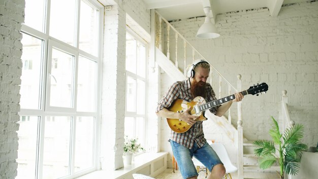 Gracioso hombre barbudo baila en la cama cantando y tocando guitarra eléctrica en el dormitorio en casa