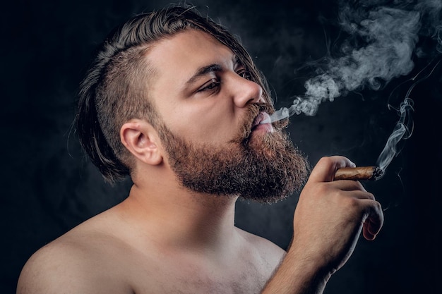 Gracioso hombre barbudo sin camisa fumando un cigarro sobre fondo gris.