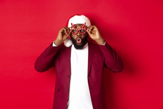 Gracioso hombre negro celebrando el año nuevo, con gafas de fiesta y gorro de Papá Noel, divirtiéndose sobre fondo rojo.