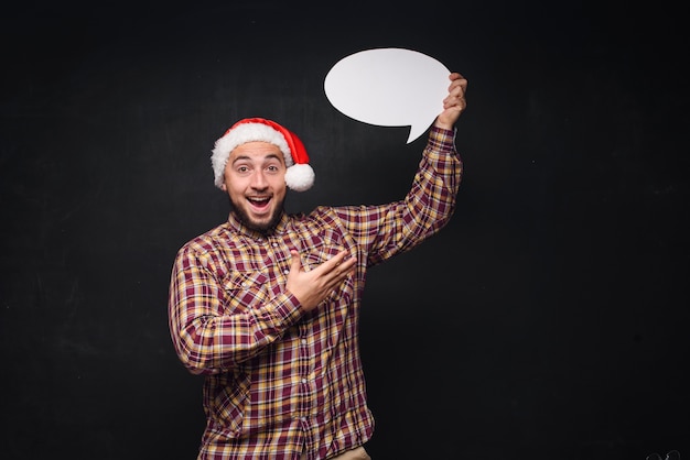 Gracioso hombre serio con sombrero rojo de Navidad Santa tiene cartón blanco vacío como blanco o simulacro con espacio de copia de texto. Fondo negro