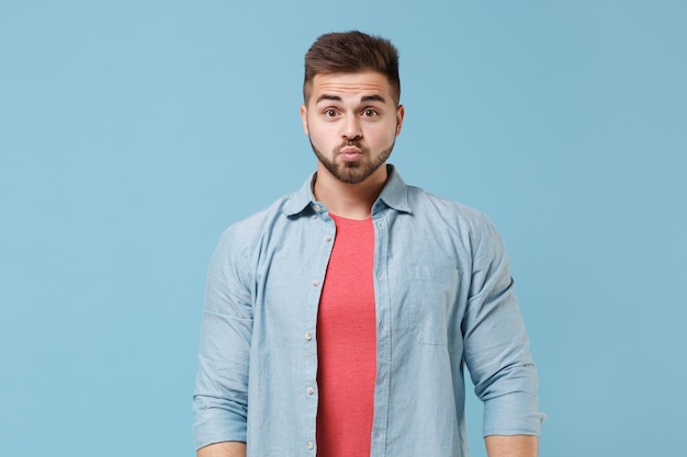 Gracioso joven barbudo de 20 años con camisa casual posando aislado en un retrato de estudio de fondo azul pastel. Emociones sinceras de la gente, concepto de estilo de vida. Simulacros de espacio de copia. Soplando los labios, haciendo cara de pato.