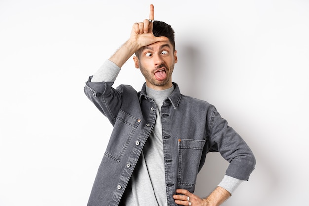 Gracioso joven haciendo muecas y mostrando la lengua con el signo de perdedor en la frente, entrecerrando los ojos, de pie sobre fondo blanco.